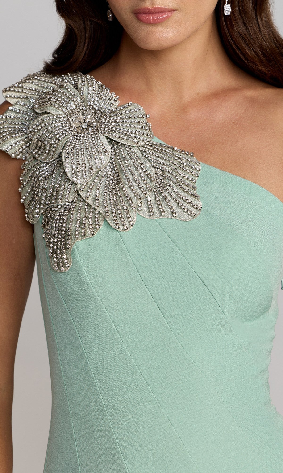 A woman wears the Mac Duggal 13221 Tea-Length Party Dress in light green crepe, showcasing a striking one-shoulder silhouette adorned with an embellished floral applique featuring bold silver beaded detailing.